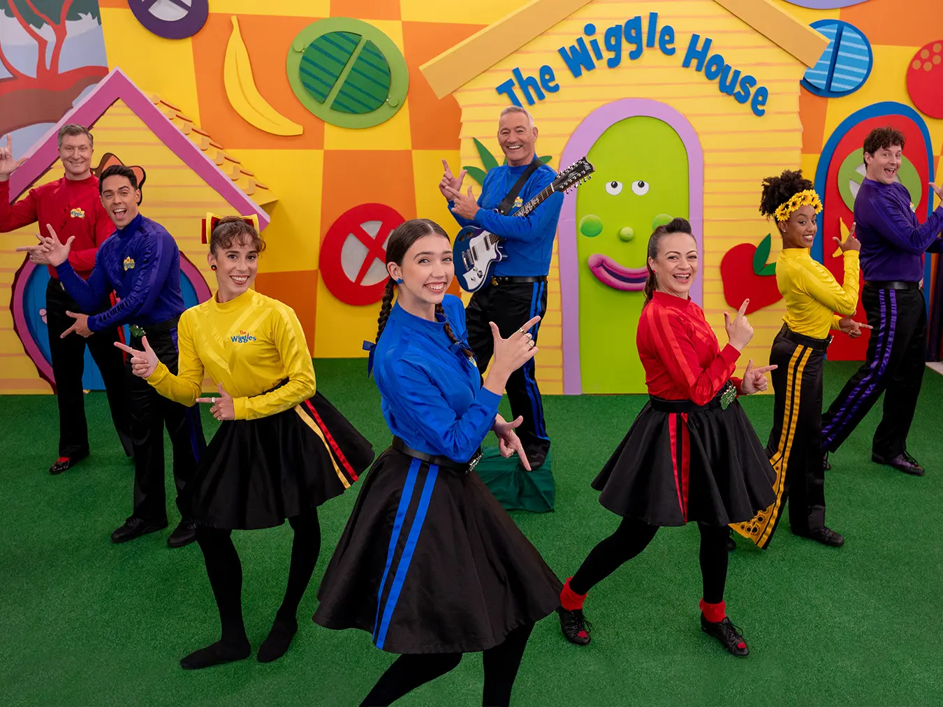 The Wiggles group, dressed in their signature bold colors, stands in front of a set in the shape of a house.