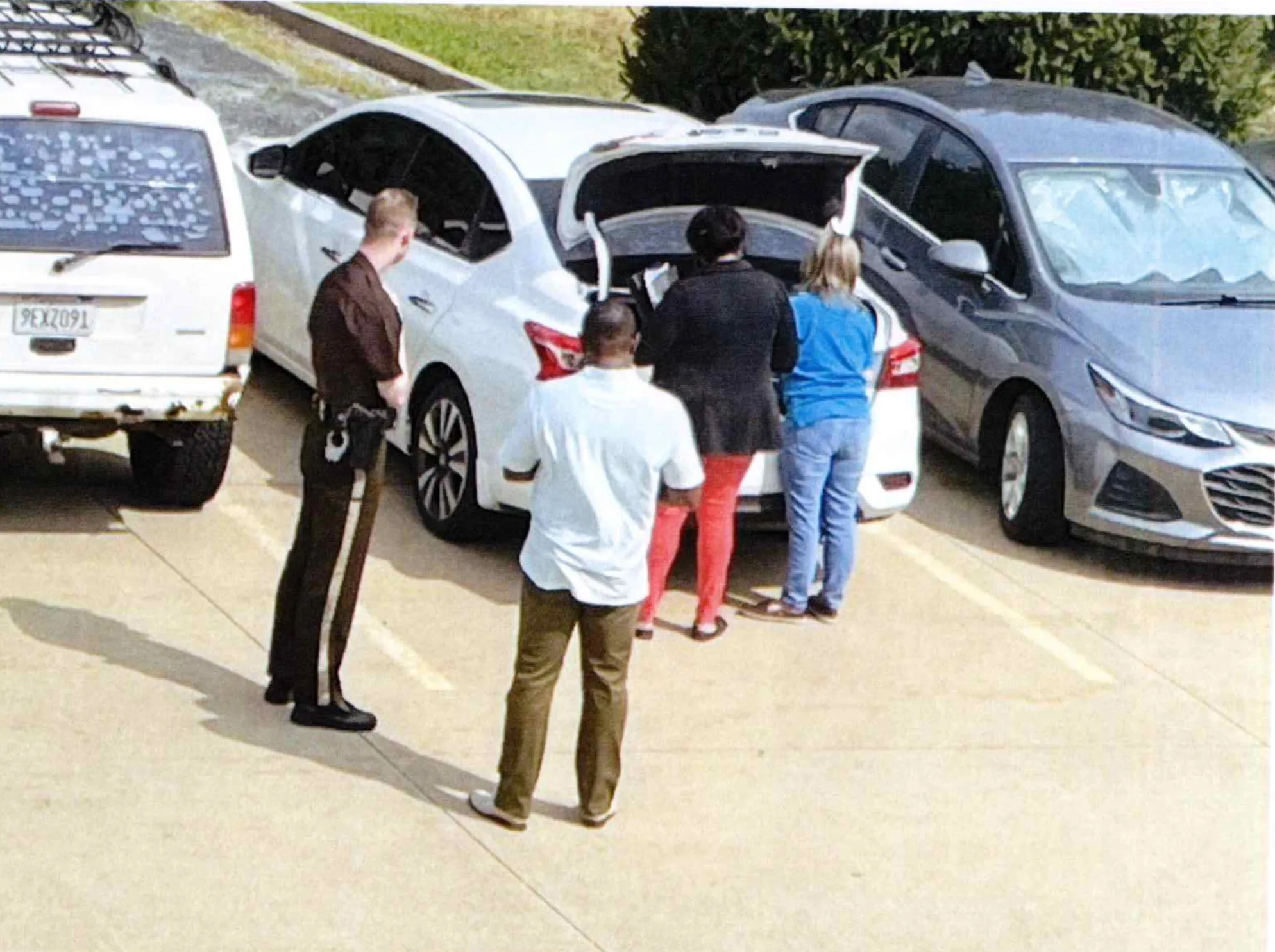 A surveillance photo shows two women looking into a car trunk while an officer and another man look on.