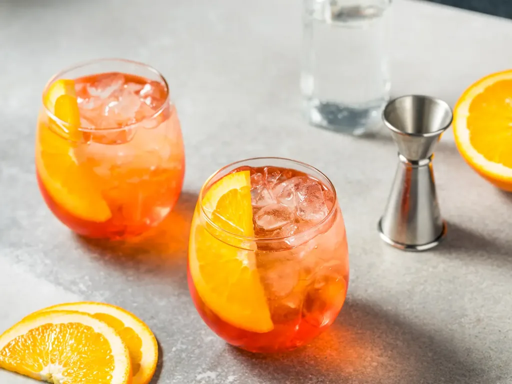 Two bright colored orange cocktails in stemless wine glasses garnished with orange slices