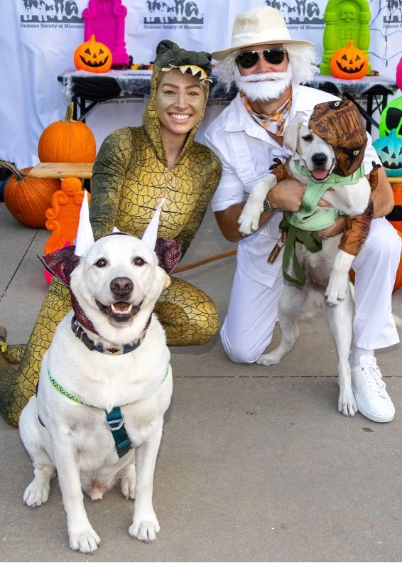 Attendees of Barktoberfest posing with their dogs in front of a step-and-repeat, all dressed in costumes.