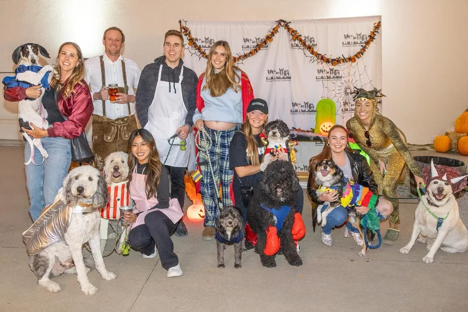 Attendees of Barktoberfest posing with their dogs in front of a step-and-repeat, all dressed in costumes.