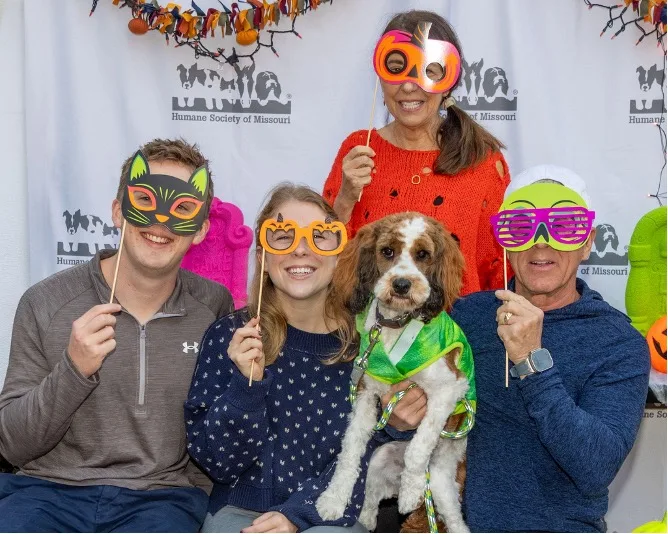 Attendees of Barktoberfest posing with their dog in front of a step-and-repeat, holding holiday eye cover holders.