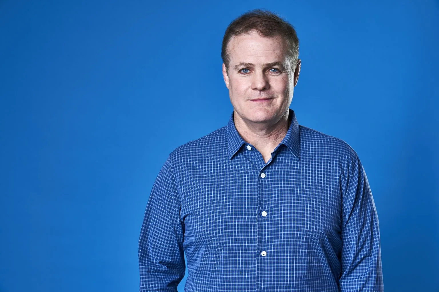Comedian Greg Warren wears a blue shirt before a blue background.