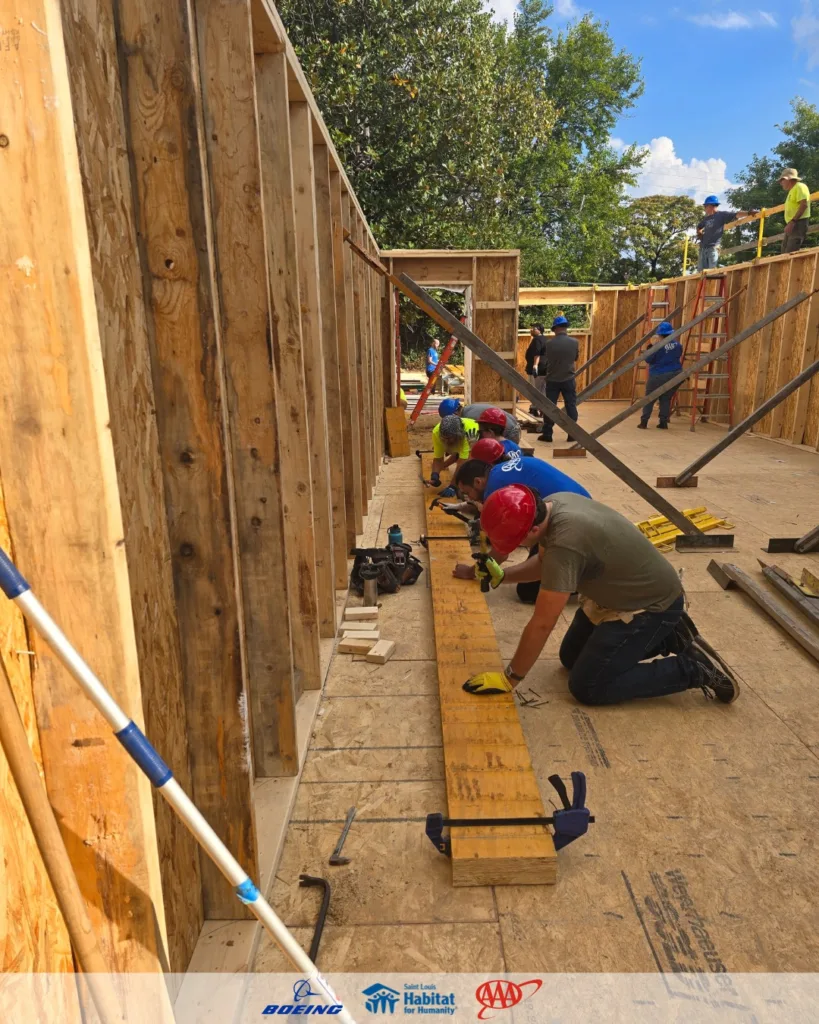 Habitat for Humanity Saint Louis volunteers in hard hats assemble wooden beams inside a partially framed home.