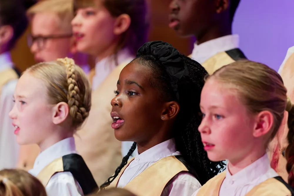 A group of young singers perform together, dressed in coordinated cream and black vests over white shirts.
