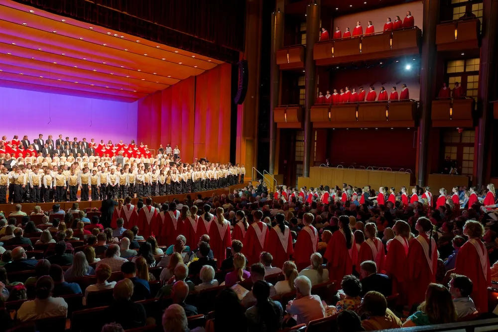 The St. Louis Children’s Choirs performs onstage in a concert hall, with audience members seated below and singers in red robes along the balconies.