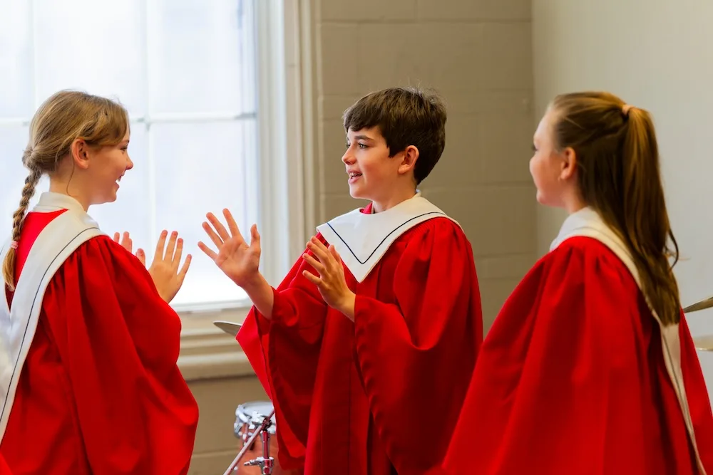 Three young choir members in red robes smile and talk backstage before a performance.