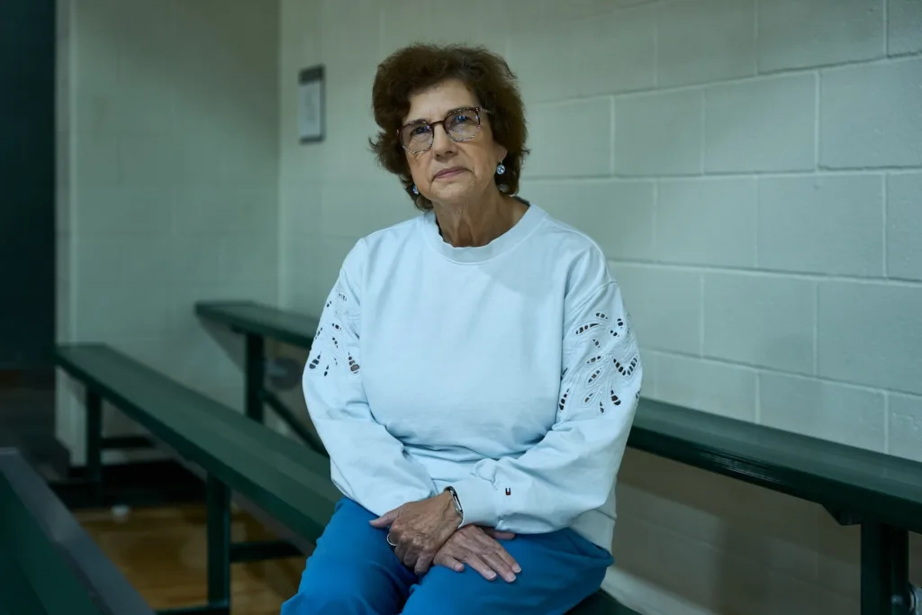 Ellen McCloskey faces the camera while sitting on the bleachers and wearing a white sweatshirt.