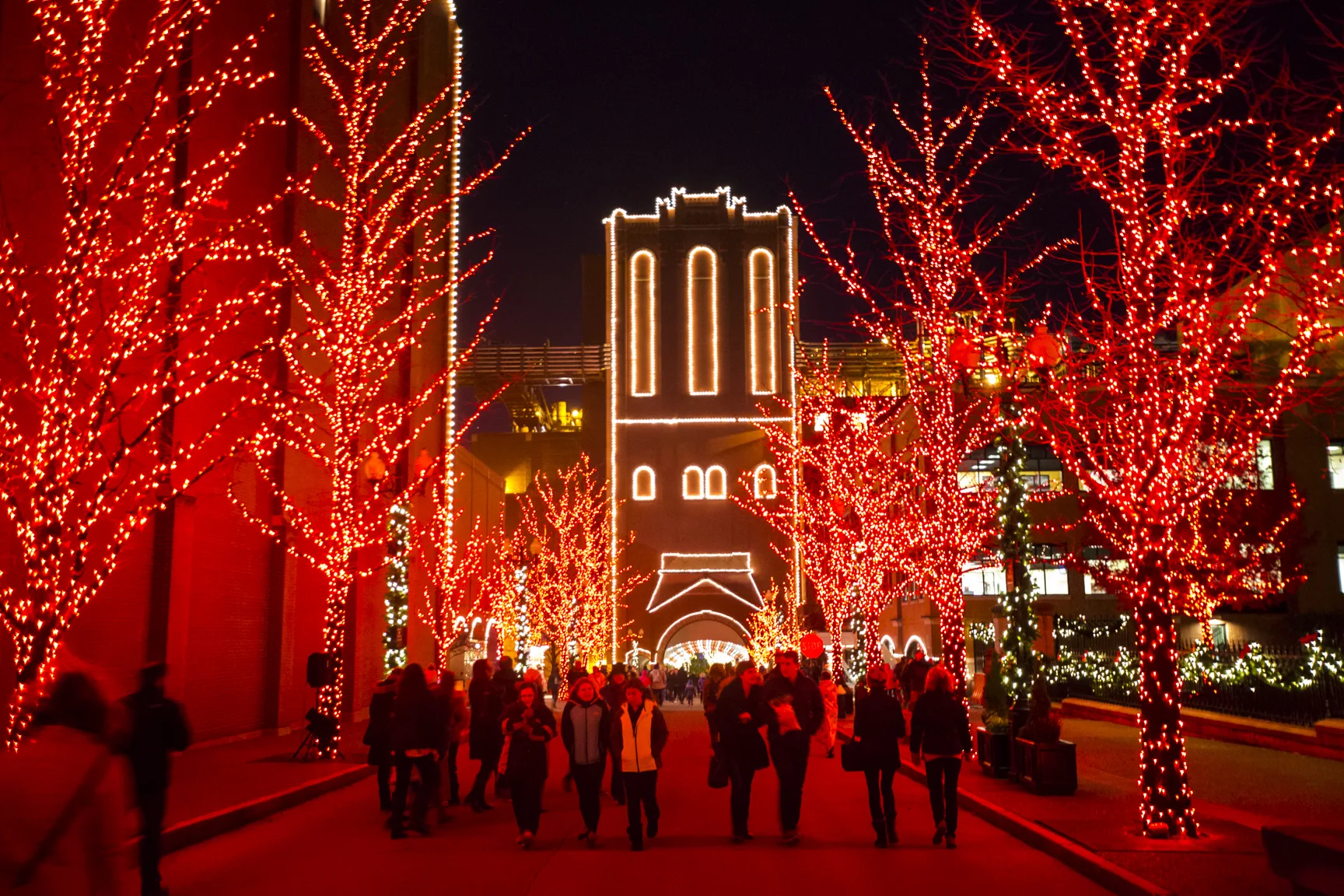 Visitors walk along a pathway lined with red-lit trees and brewery buildings accented with holiday lights.