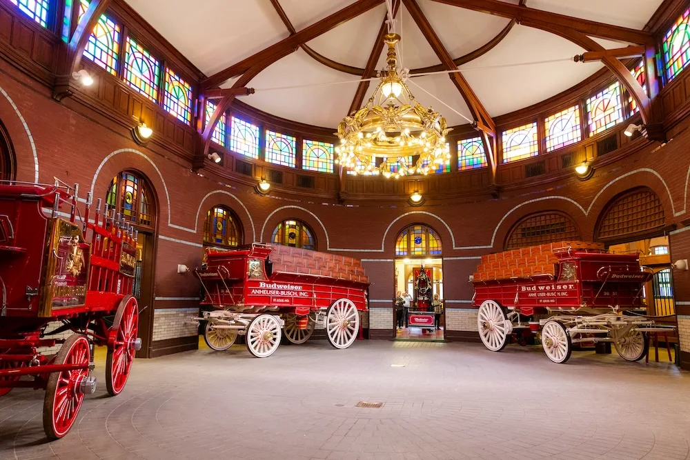 An indoor stable with stained-glass windows and historic red Budweiser wagons on display beneath a large chandelier.