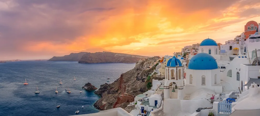 White buildings with blue domes overlook the sea in Santorini, Greece, at sunset.