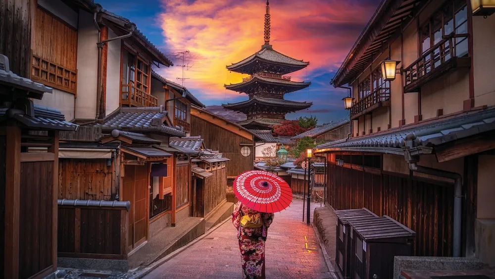 A person in traditional dress walks through a historic street in Japan at dusk.