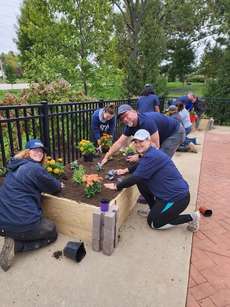 Volunteers planting flowers in a raised garden bed outside near a black metal fence.