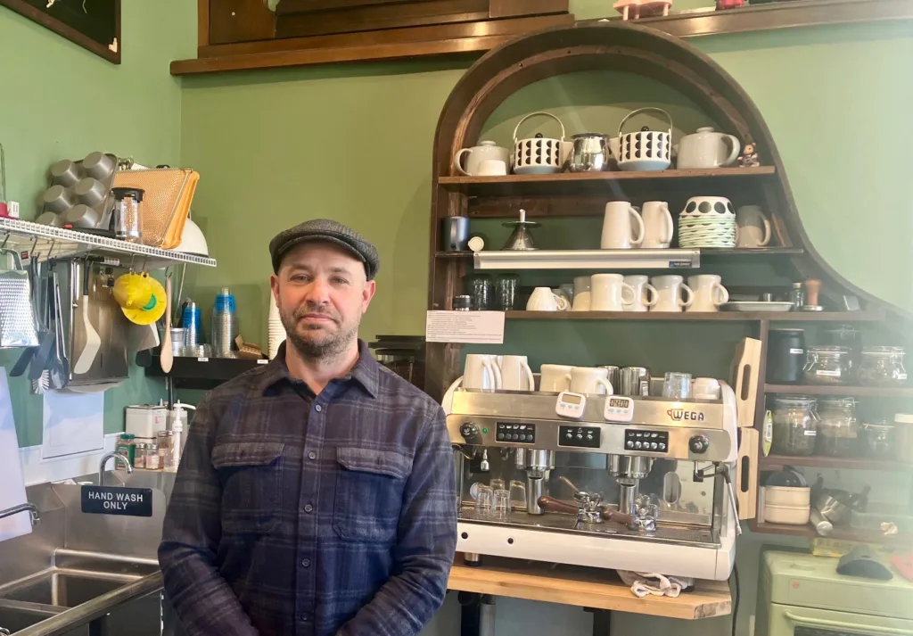 A man in a cap stands before a row of coffee mugs.