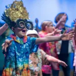 Students wearing feathered headdresses and oversized goggles perform with outstretched arms alongside classmates in tropical outfits.