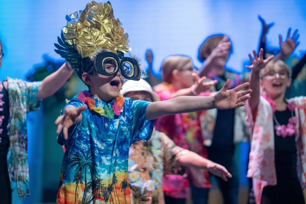 Students wearing feathered headdresses and oversized goggles perform with outstretched arms alongside classmates in tropical outfits.