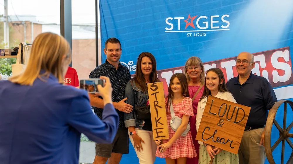 A family poses for a photo in front of a STAGES St. Louis backdrop for the Newsies production at the Kirkwood Performing Arts Center.
