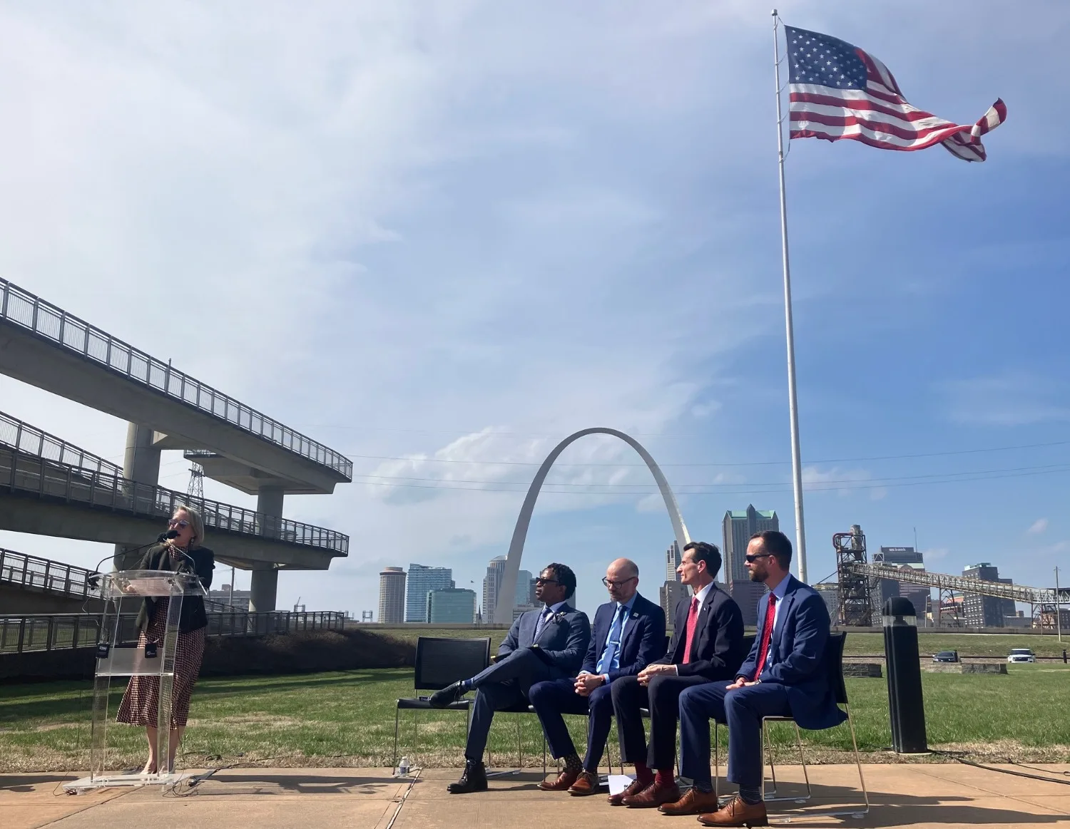 With the Arch and the American flag in the background, a congresswoman speaks while four officials watch from a bench.