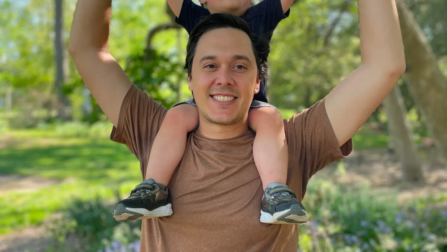 A man faces the camera with his child on his shoulders; the child's face cannot be seen.