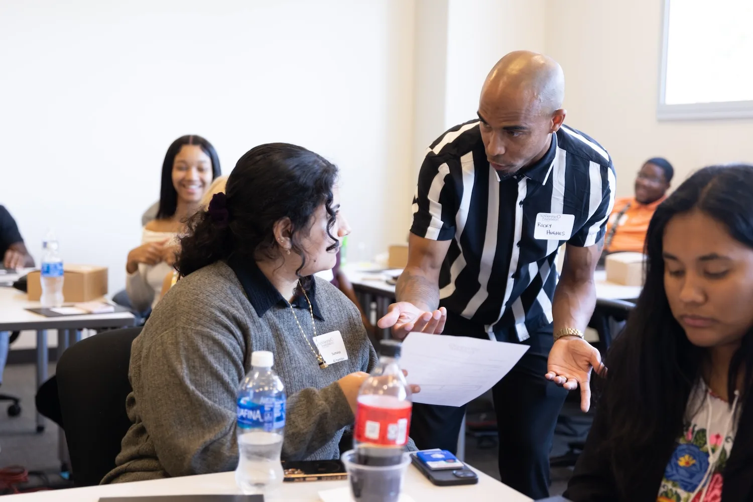 A man in a striped shirt leans down to talk with a student seated at a desk.