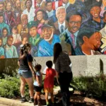 Visitors admire a large outdoor mural filled with vibrant portraits representing culture, history, and community heritage.