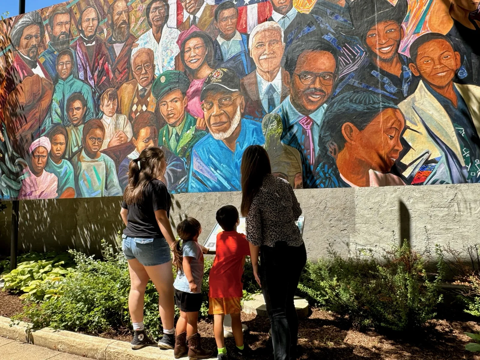 Visitors admire a large outdoor mural filled with vibrant portraits representing culture, history, and community heritage.
