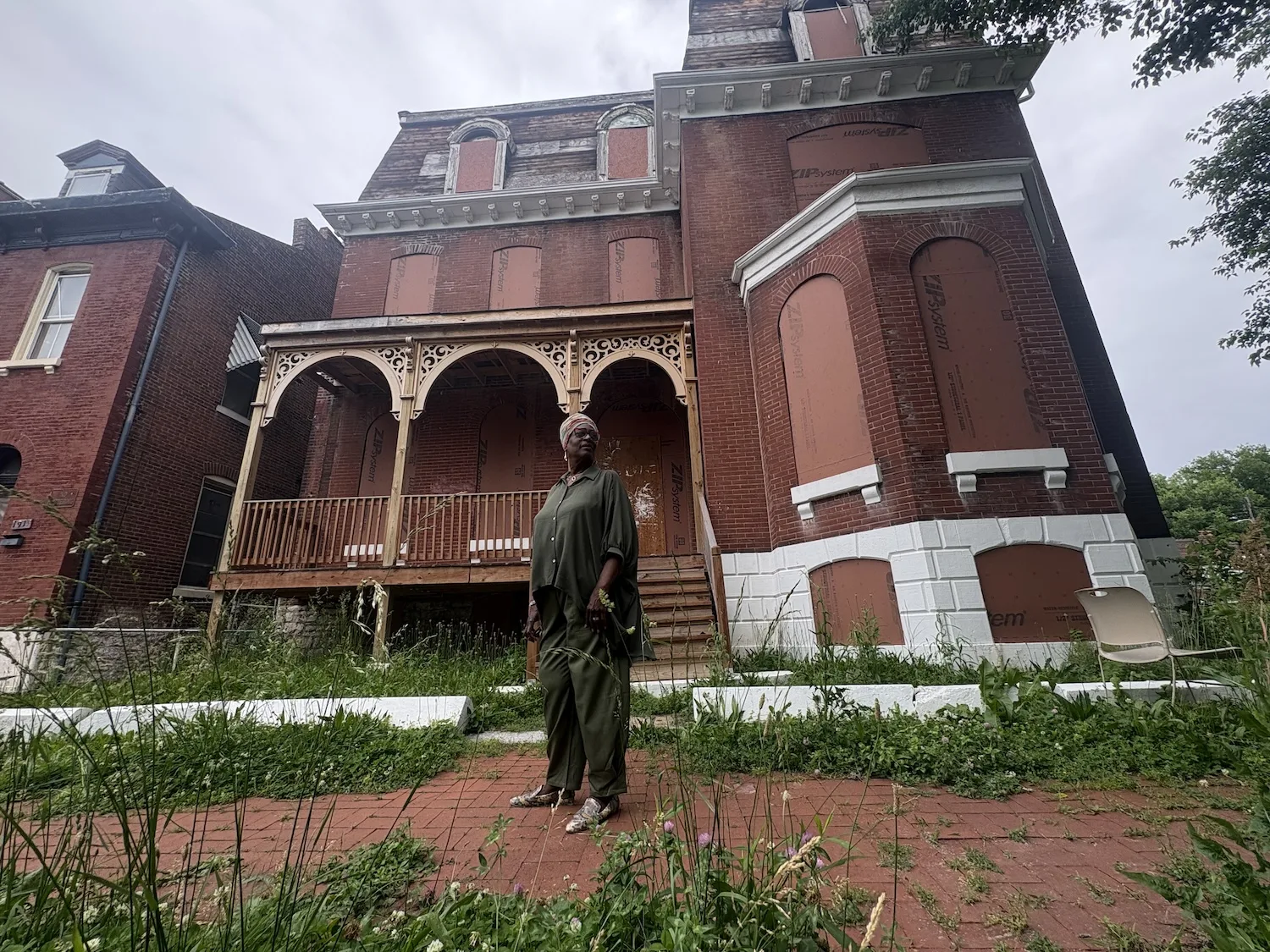 A woman stands before a boarded up Victorian-era home.