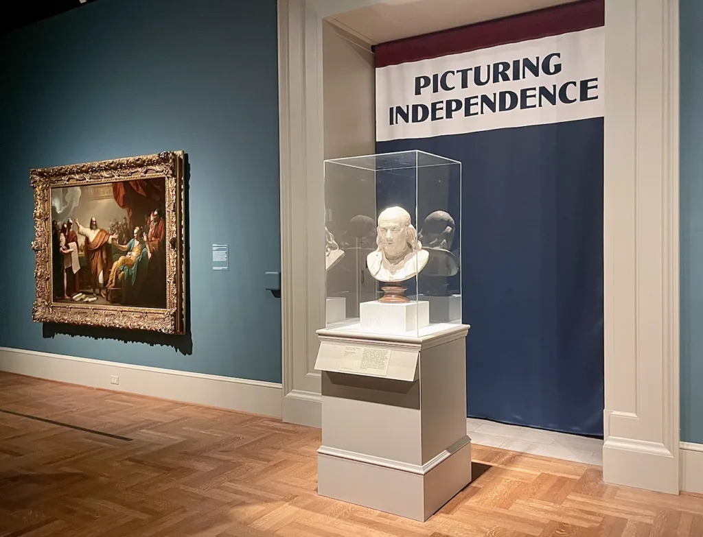 The "Revolution" section of 'Picturing Independence,' featuring Jean-Antoine Houdon's bust of Benjamin Franklin and Benjamin West's "Daniel Interpreting to Belshazzar the Writing on the Wall."