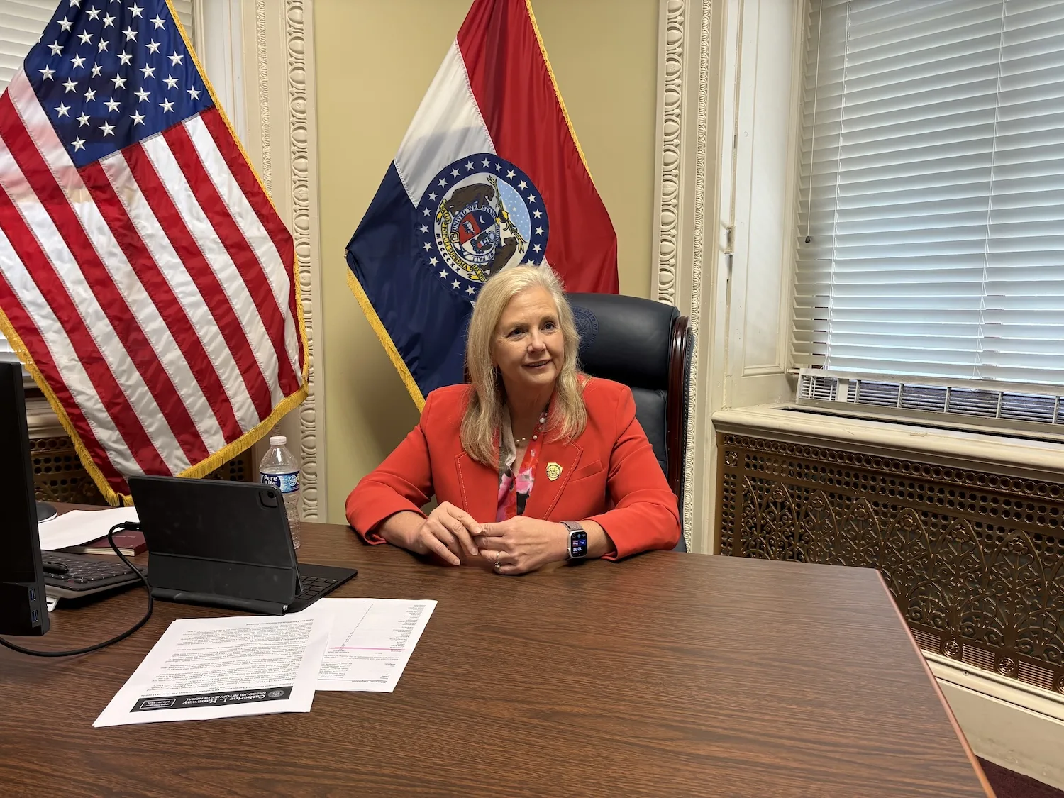 A woman in a red suit sits at a desk with the American and Missouri flags behind her.