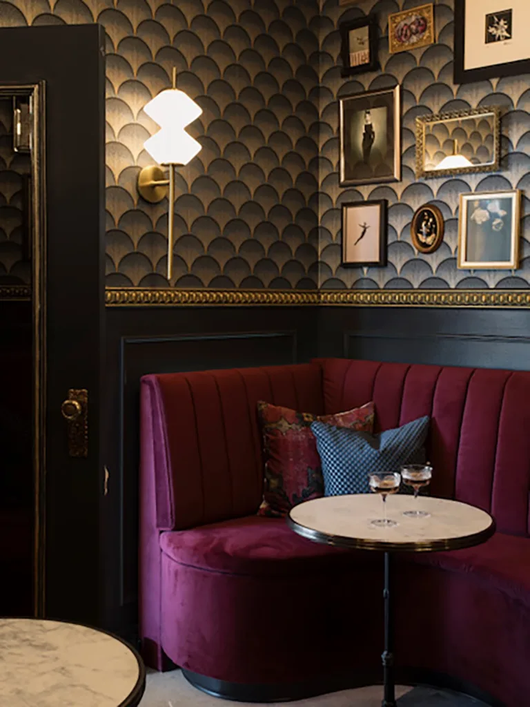 A dimly-lit speakeasy lounge with opulent wallpaper, red velvet seating and two cocktails on a small white table.