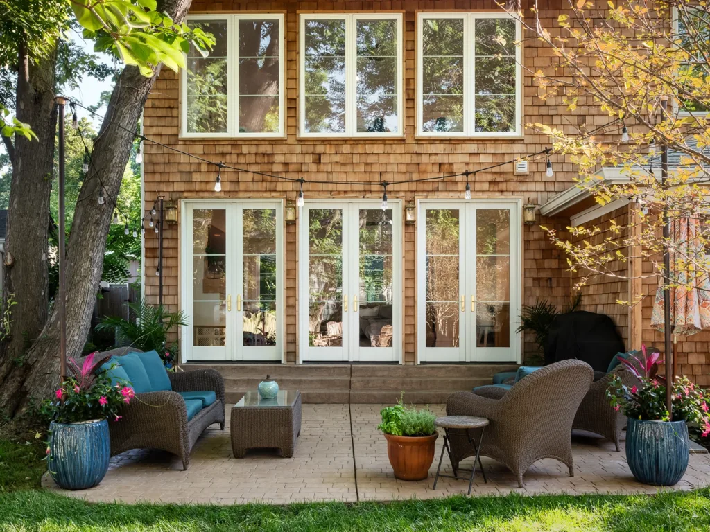 The backyard exterior view of a newly renovated, historic home in Webster Groves with warm-toned brick, large windows and French doors leading to a cozy patio area with a couch, two chairs, a coffee table, string lights and potted plants.