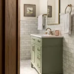 A warm and bright bathroom with cream-colored walls, gray and white stone tiling, a sage green vanity, curved vanity mirror and gold hardware accents.