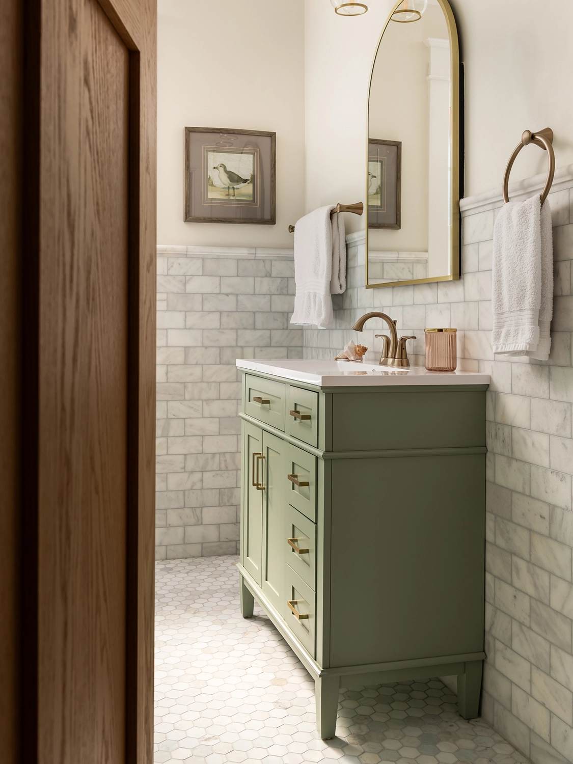 A warm and bright bathroom with cream-colored walls, gray and white stone tiling, a sage green vanity, curved vanity mirror and gold hardware accents.
