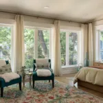 Primary bedroom suite in a newly renovated historic home with almost-floor-to-ceiling windows, a floral area rug, two accent chairs and a bed.