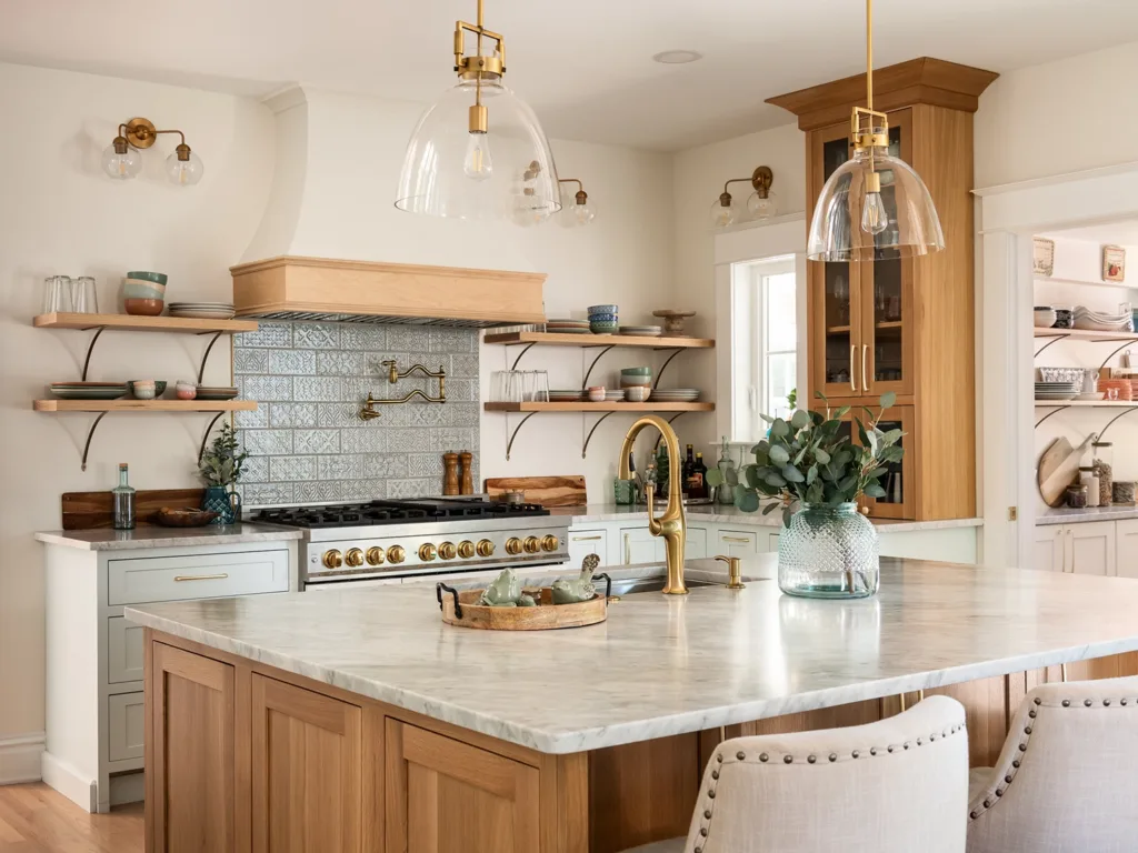 The bright and airy newly renovated kitchen of a historic home in Webster Groves, featuring pale blue cabinets and a large light-toned wooden island with light gray marble countertops, open shelving and gold accents.