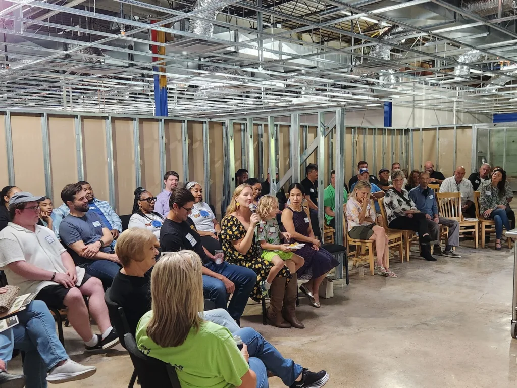 People sit in chairs around an unfinished room, as one woman holds a microphone.