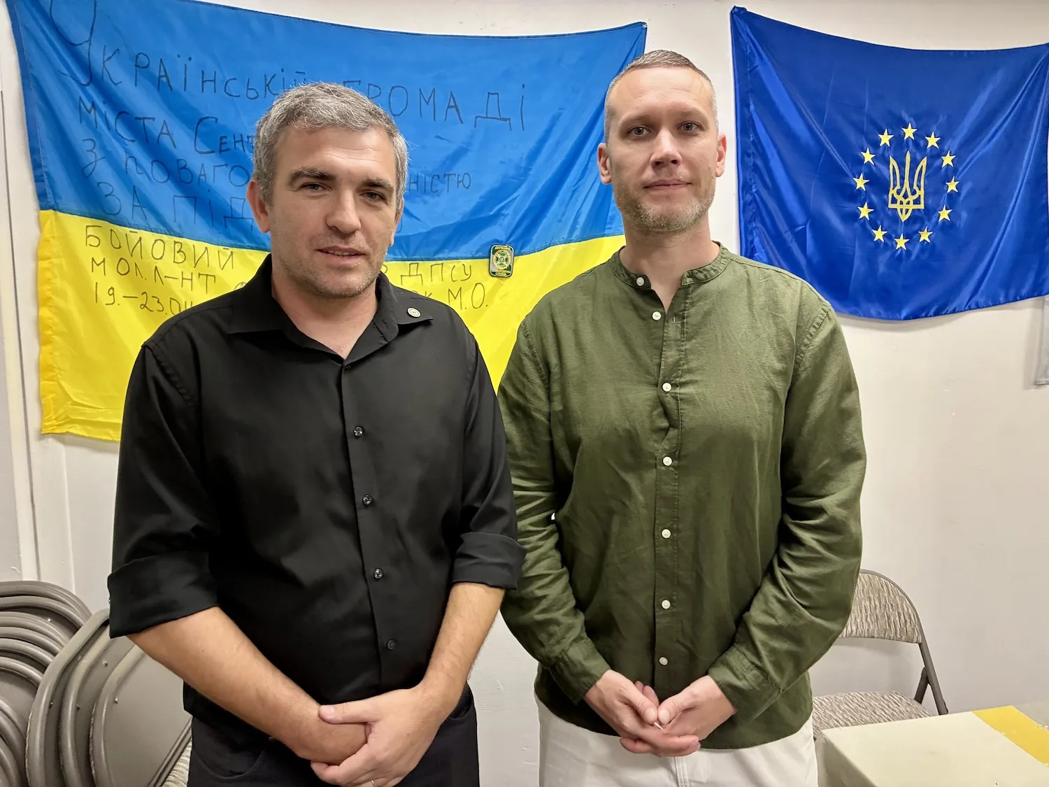 Two men stand in front of an Ukrainian flag.